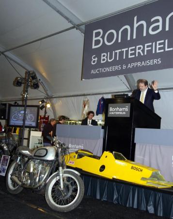 bud ekins frenzy at petersen museum auction, Bonhams CEO and auctioneer Malcolm Barber gives notice for final bids on a legendary 1950s Norton Manx racer Though not running and certainly not restored it was expected to bring 10 15 000 but brought 30 000 The yellow body seen in the background belongs to a CART IndyCar 800 hp racer that sold for about 20 000