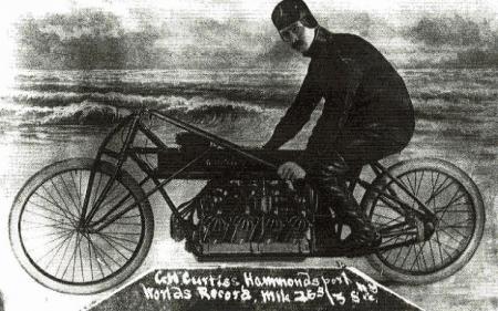 motorcycle history part 2, Glenn Curtiss set a world speed record in 1907 aboard this self built V8 powered contraption Wearing a leather cap a well manicured moustache and a steely gaze he stares out of this time worn black and white postcard like photo In the background frothy waves splash upon the hard packed sands upon which his experimental hot rod motorcycle would transport him into the history books as The Fastest Man in the World